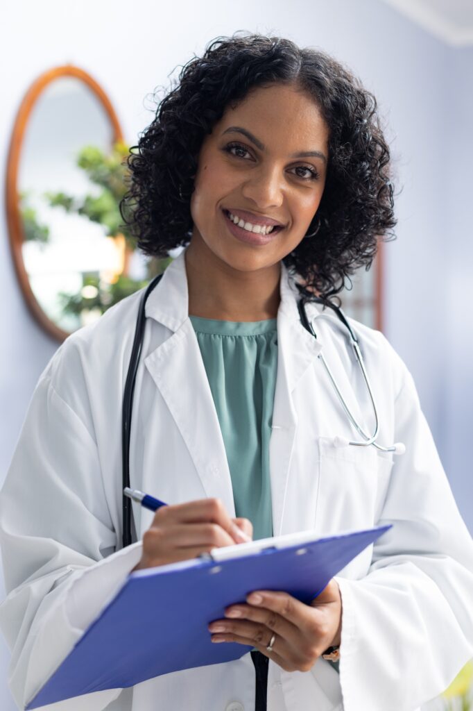 Portrait of happy biracial female doctor holding clipboard and smiling at doctor's office
