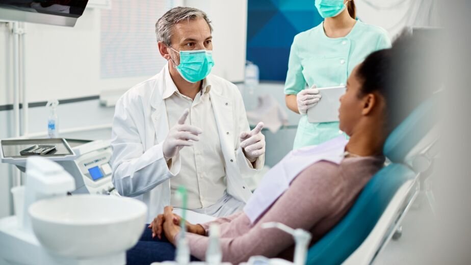 Mature dentist with face mask talking to black female patient at dentist's office.