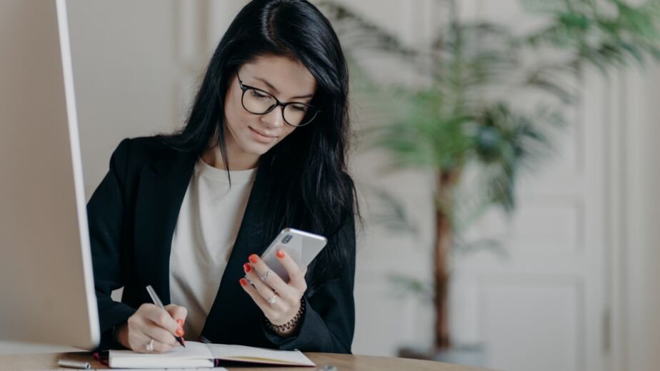 Woman with dark hair, concentrated into mobile phone, searches necessary information in internet